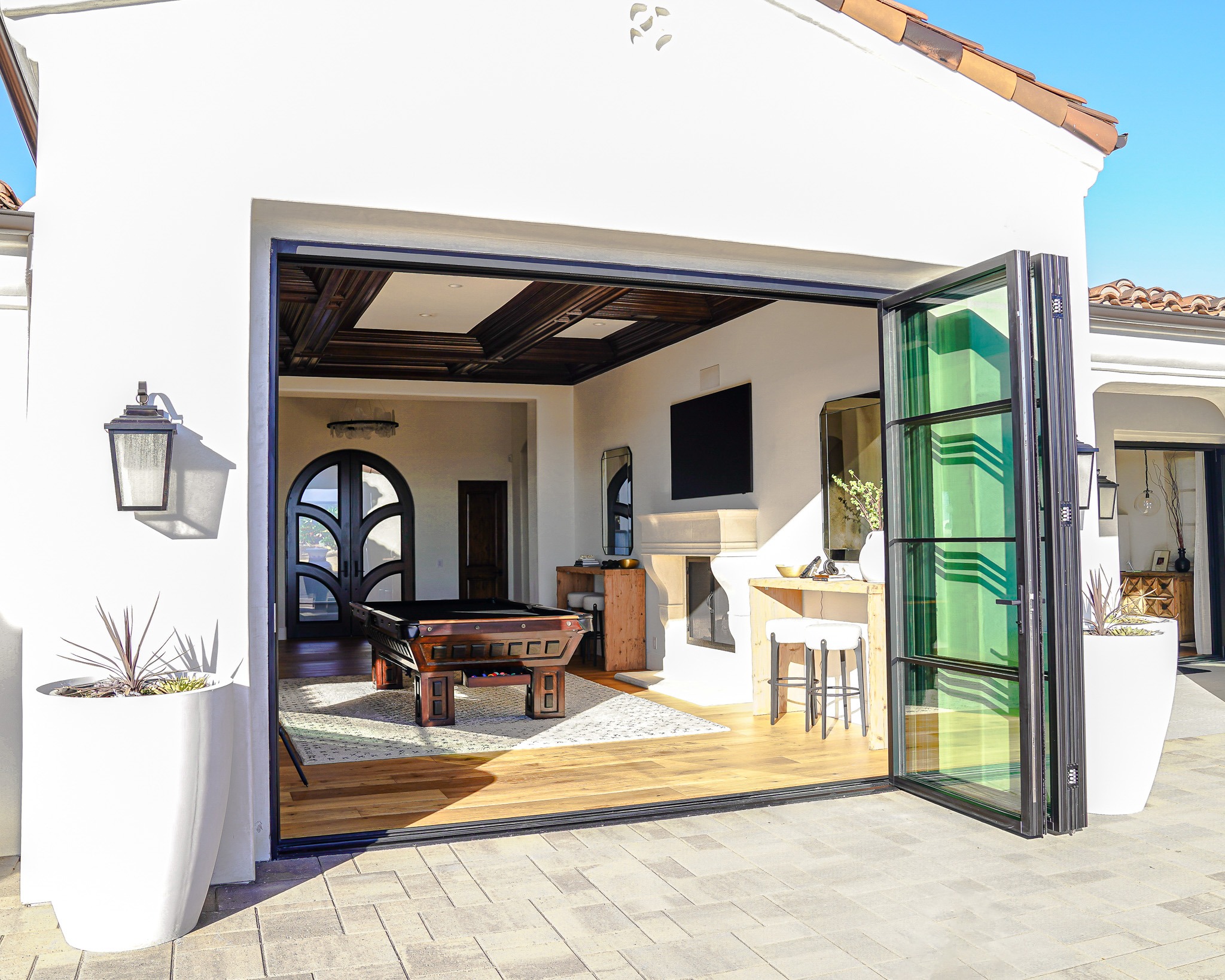 Modern home patio with large sliding glass doors open to a stylish interior living space, featuring a kitchen island, wood-beamed ceiling, and furnishings.